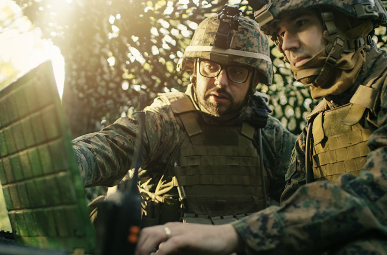 Two men in military uniforms outdoors looking at a laptop